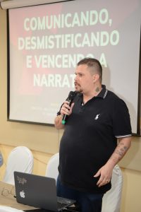 Fotografia de Marcos Paulo, um dos palestrantes do Seminário. No fundo, projeção de tela com o texto Comunicação, Desmistificando e Vencendo a Narrativa.